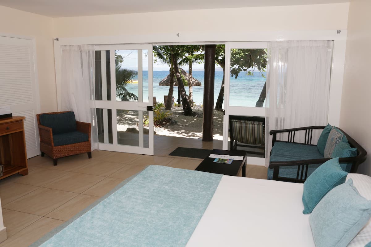 Une chambre avec un lit, deux chaises, une petite table et une vue sur la plage, accessible par une porte coulissante.