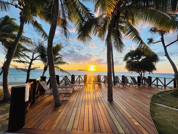 Une terrasse en bois avec des chaises longues et une rambarde d’où l’on peut admirer le coucher de soleil sur l’océan.