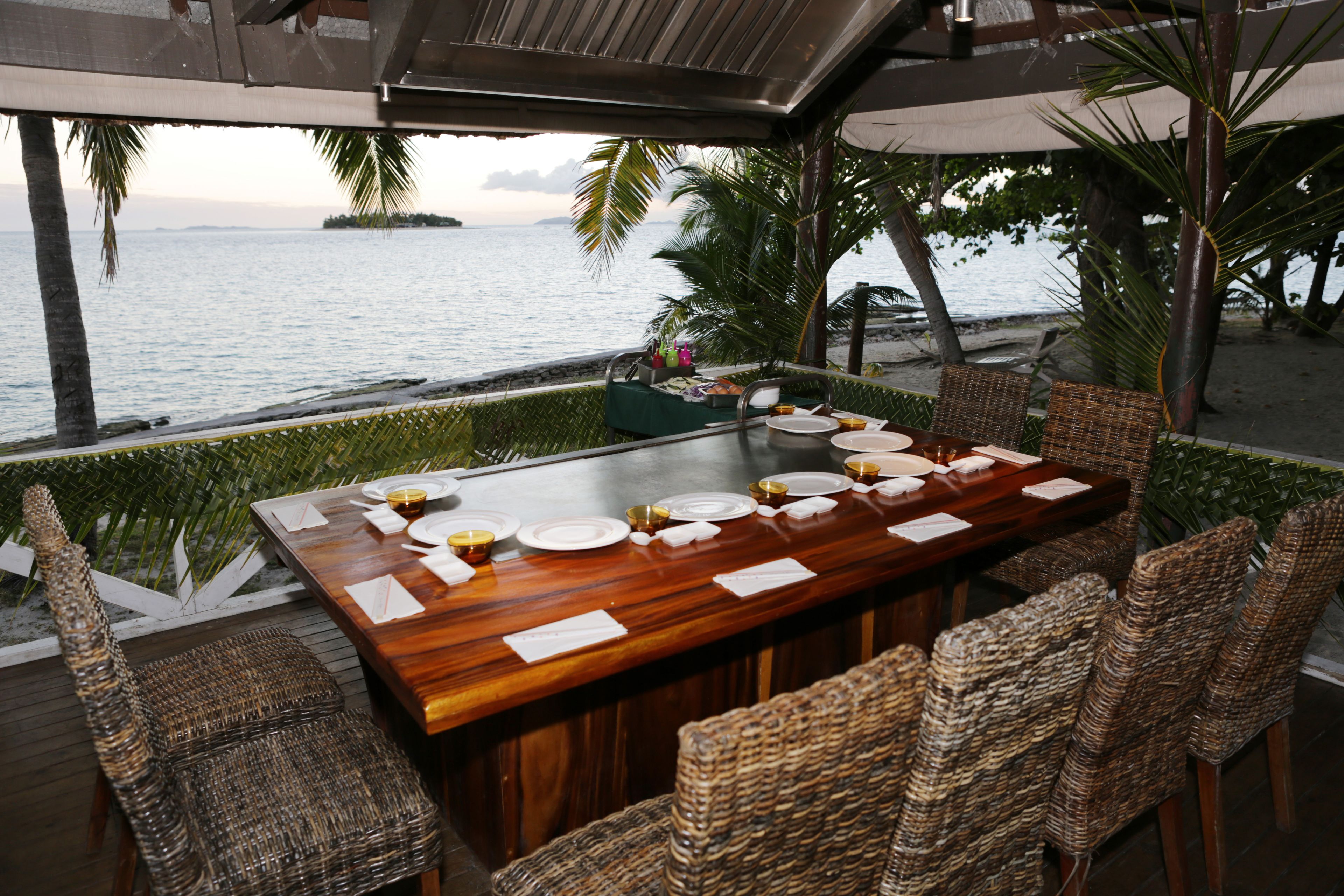 Uma sala de jantar com mesa de madeira posta para seis, rodeada por cadeiras de vime, com vista para uma praia com palmeiras e o mar.