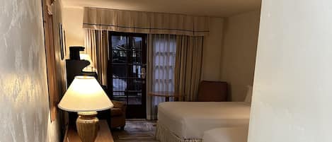 Habitación de hotel con cama, mesita de noche con lámpara, mesita auxiliar de madera y vistas a través de un ventanal.