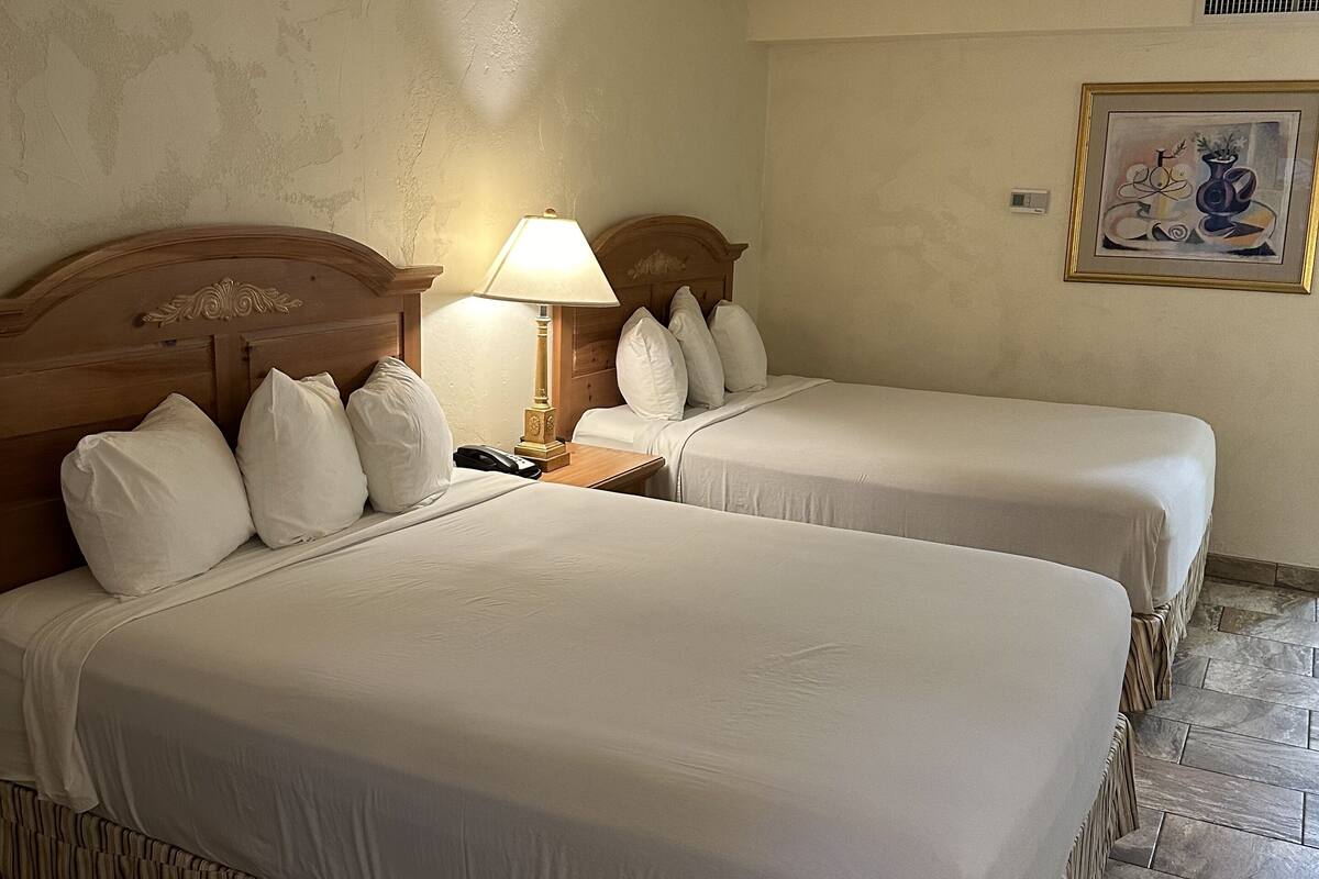 A hotel room with two beds, a nightstand, a lamp, a framed artwork, and a tiled floor.