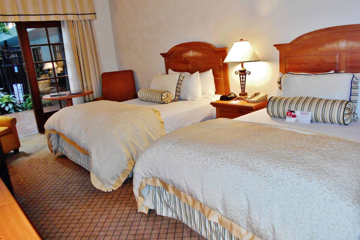 A hotel room with two beds, a wooden headboard, a bedside table with a lamp, a chair, and a view of the outdoors.