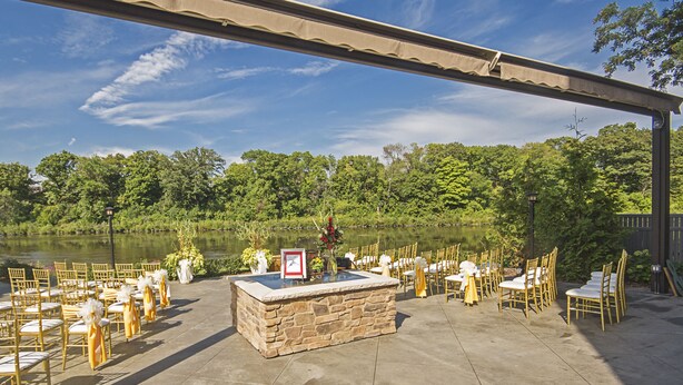 An outdoor wedding setup with stone tables and chairs, a view of a lake, and a covered area with a pergola. - Holiday Inn Milwaukee Riverfront by IHG (Milwaukee)