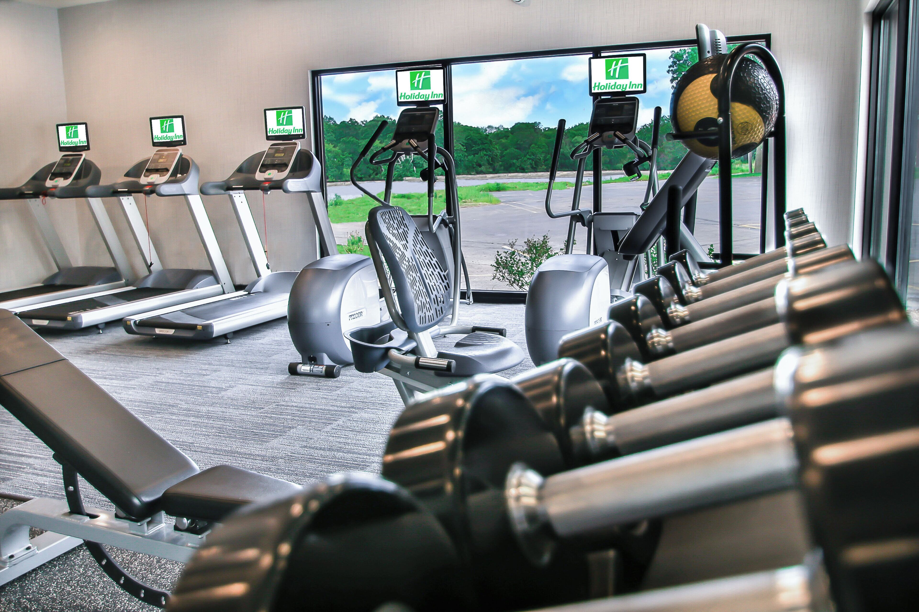 A gym with treadmills, elliptical machines, and a rowing machine, with a view of a parking lot and greenery outside.
