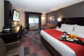Une chambre d’hôtel comprenant un lit, un bureau avec un ordinateur, un canapé, une télévision et un plateau de petit-déjeuner avec du café, du jus de fruits et des viennoiseries.