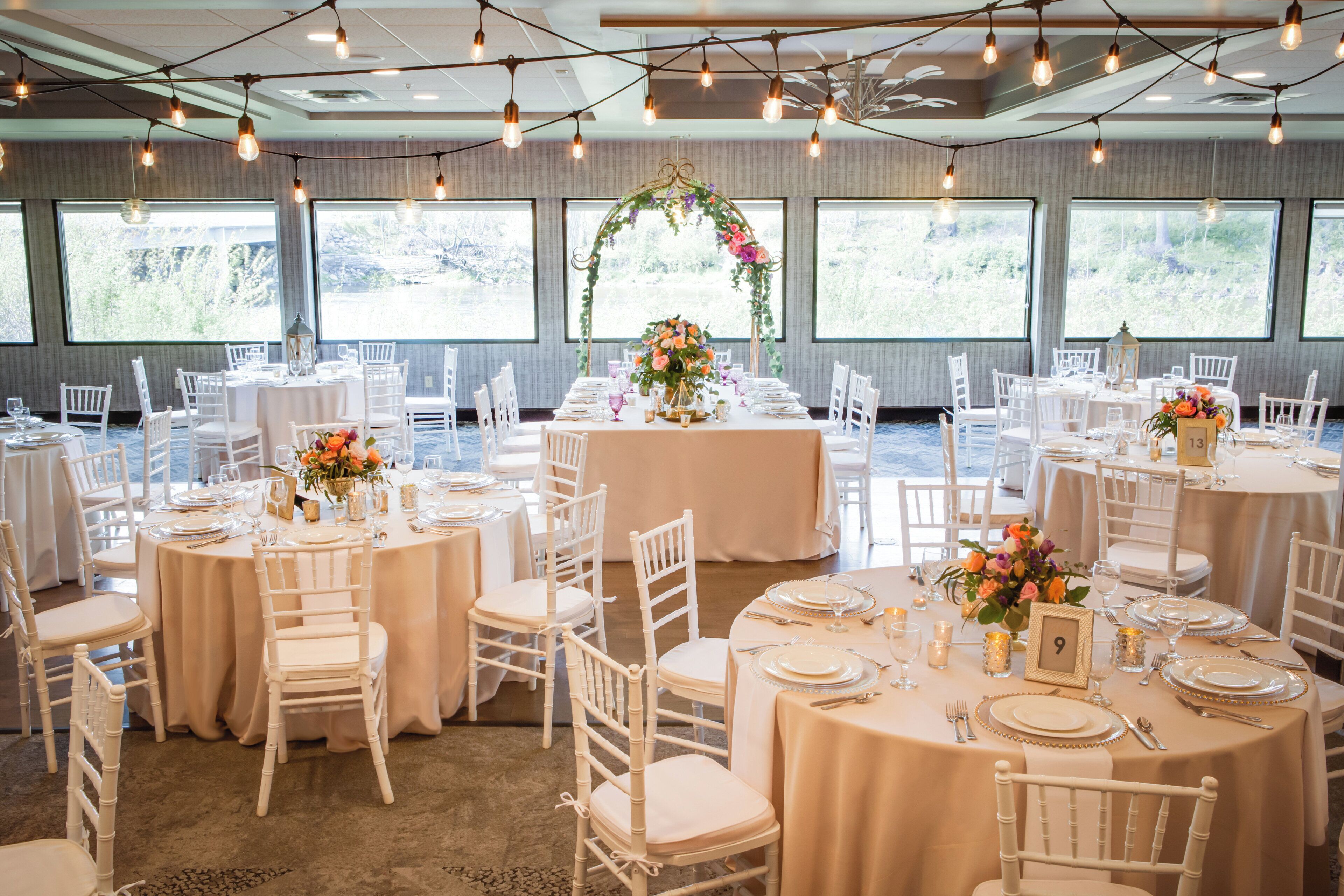 Un salón de banquetes con mesas redondas, sillas blancas y centros de mesa florales.