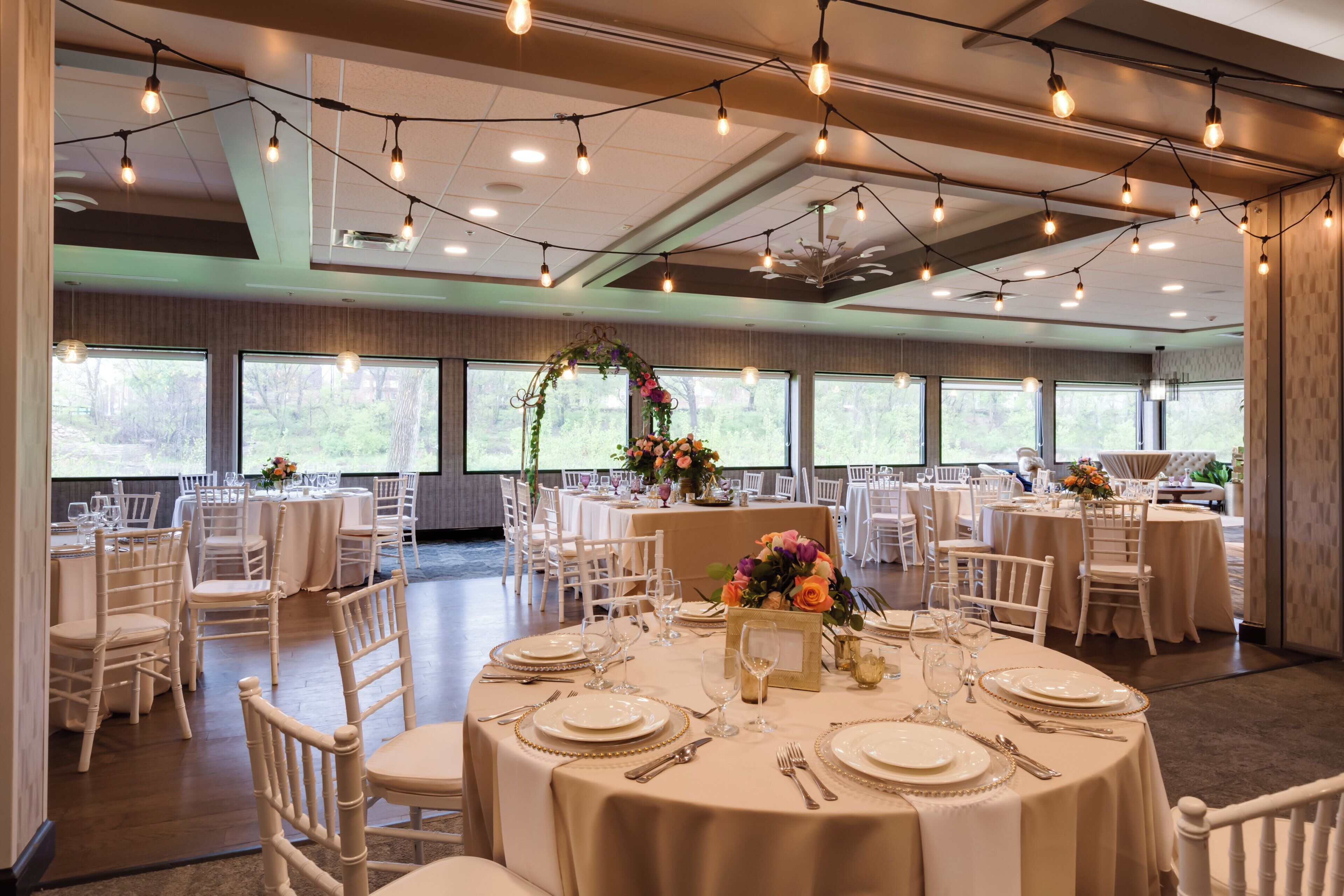 A banquet hall with round tables set for dining, large windows, and string lights.