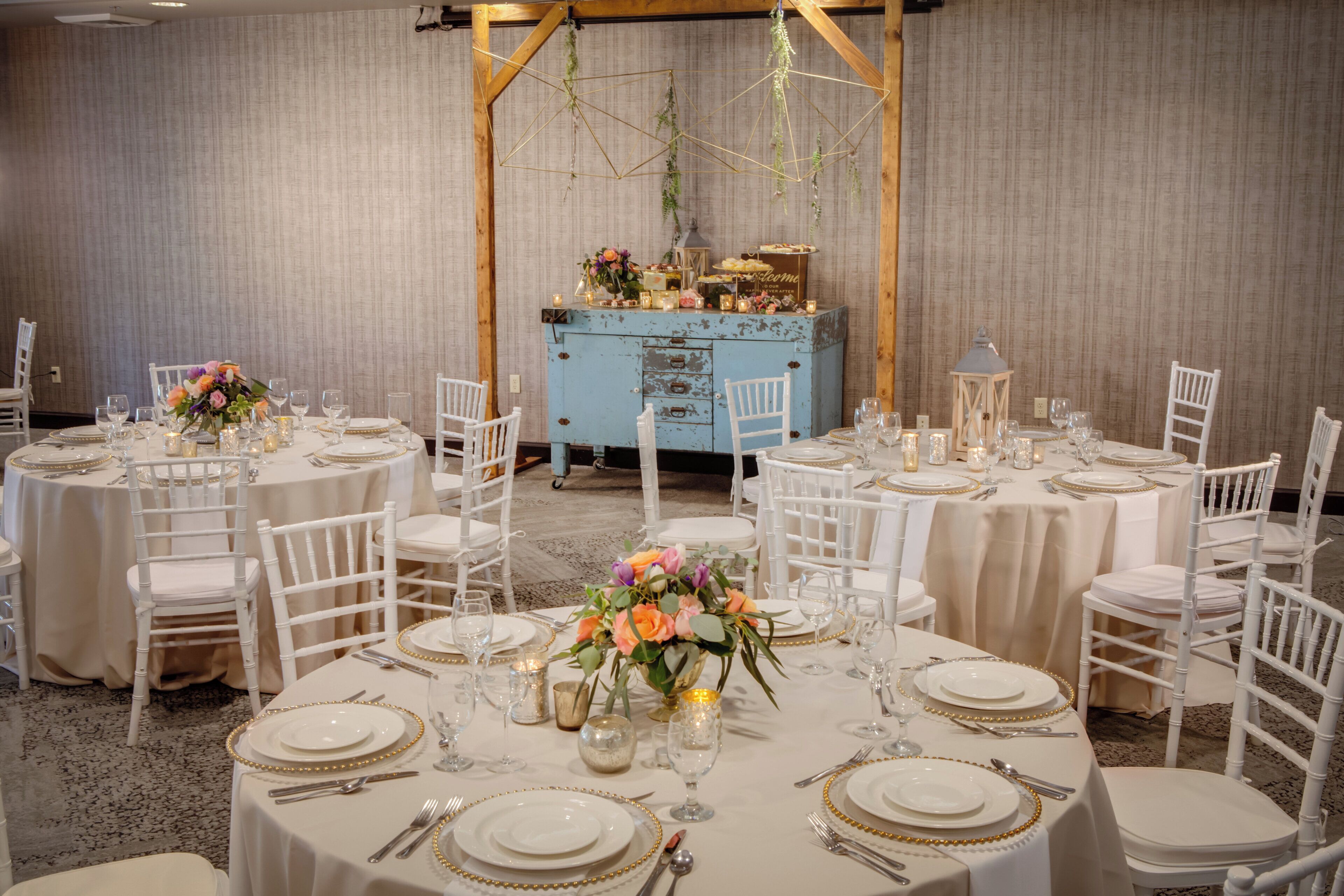 Un salón de banquetes con mesas redondas dispuestas para comer, sillas blancas y un aparador azul al fondo.