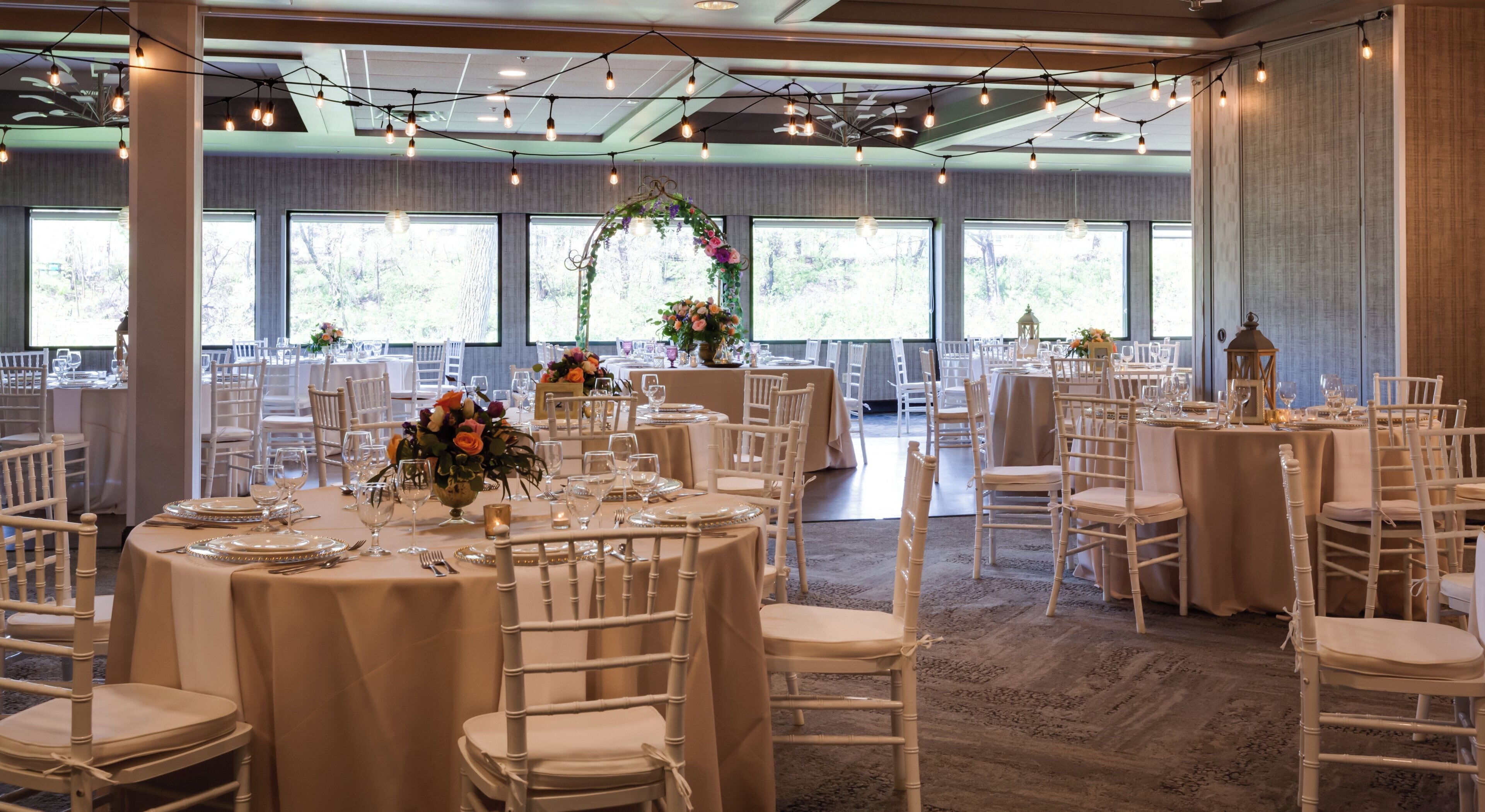 Une salle de banquet avec des tables rondes, des chaises blanches et des guirlandes lumineuses.