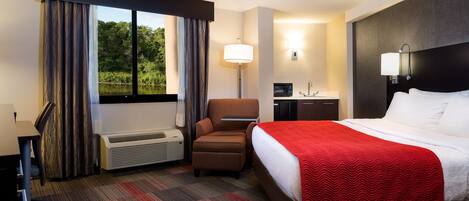 Habitación de hotel con una cama grande, un escritorio con lámpara, una silla, una mesita y una ventana con cortinas.