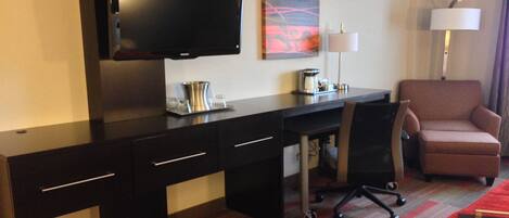 A hotel room with a flat-screen TV mounted on the wall, a desk with a chair, a coffee maker, and a lamp.