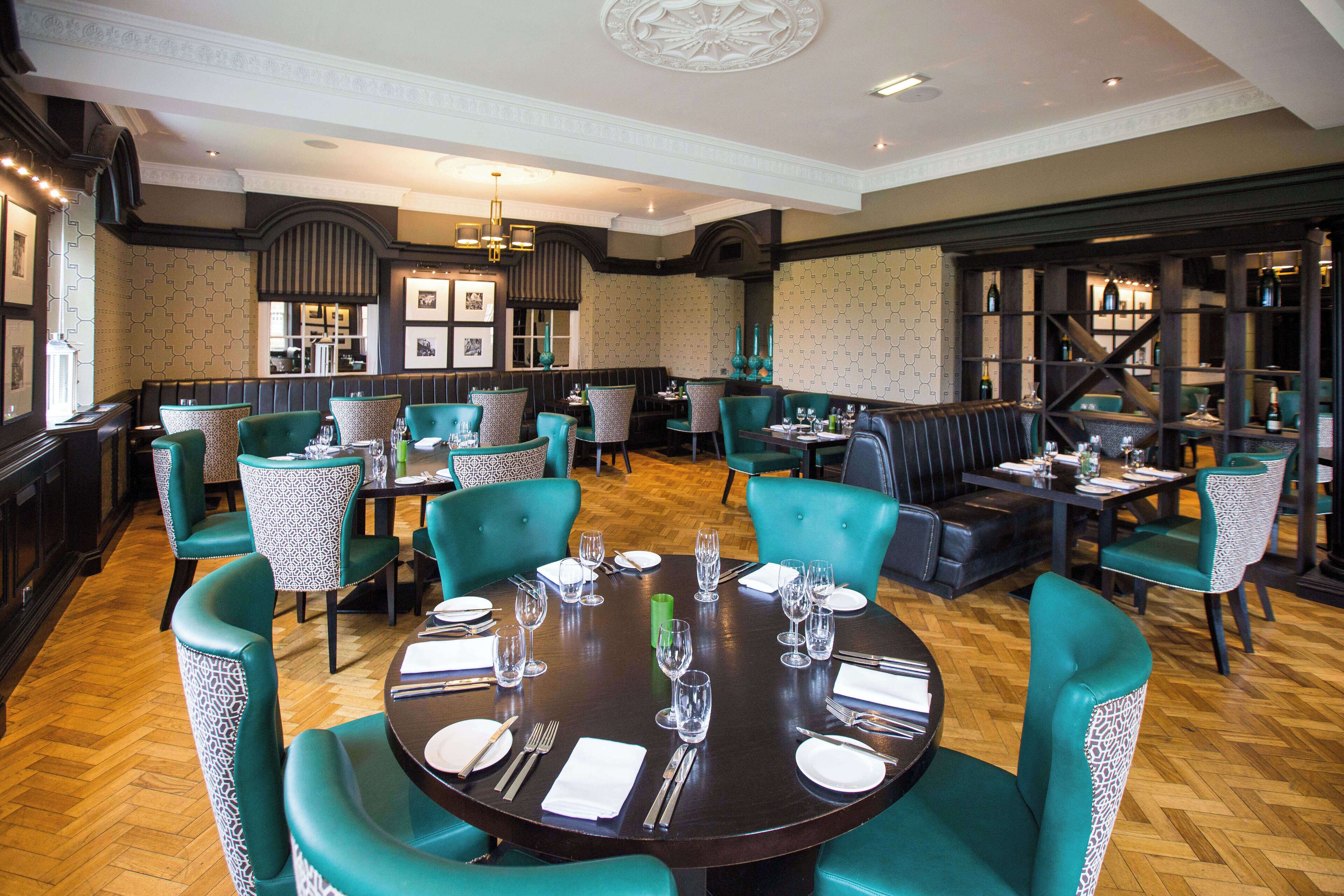 A restaurant with round tables set for dining, teal chairs, and a bar area with stools.
