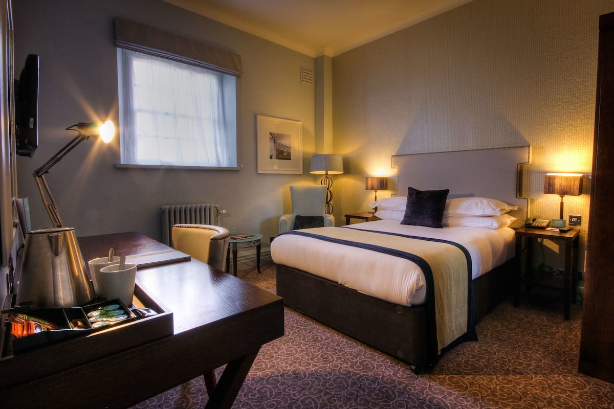 A hotel room with a large bed, bedside tables, a desk with a coffee maker, a chair, and a window with curtains.