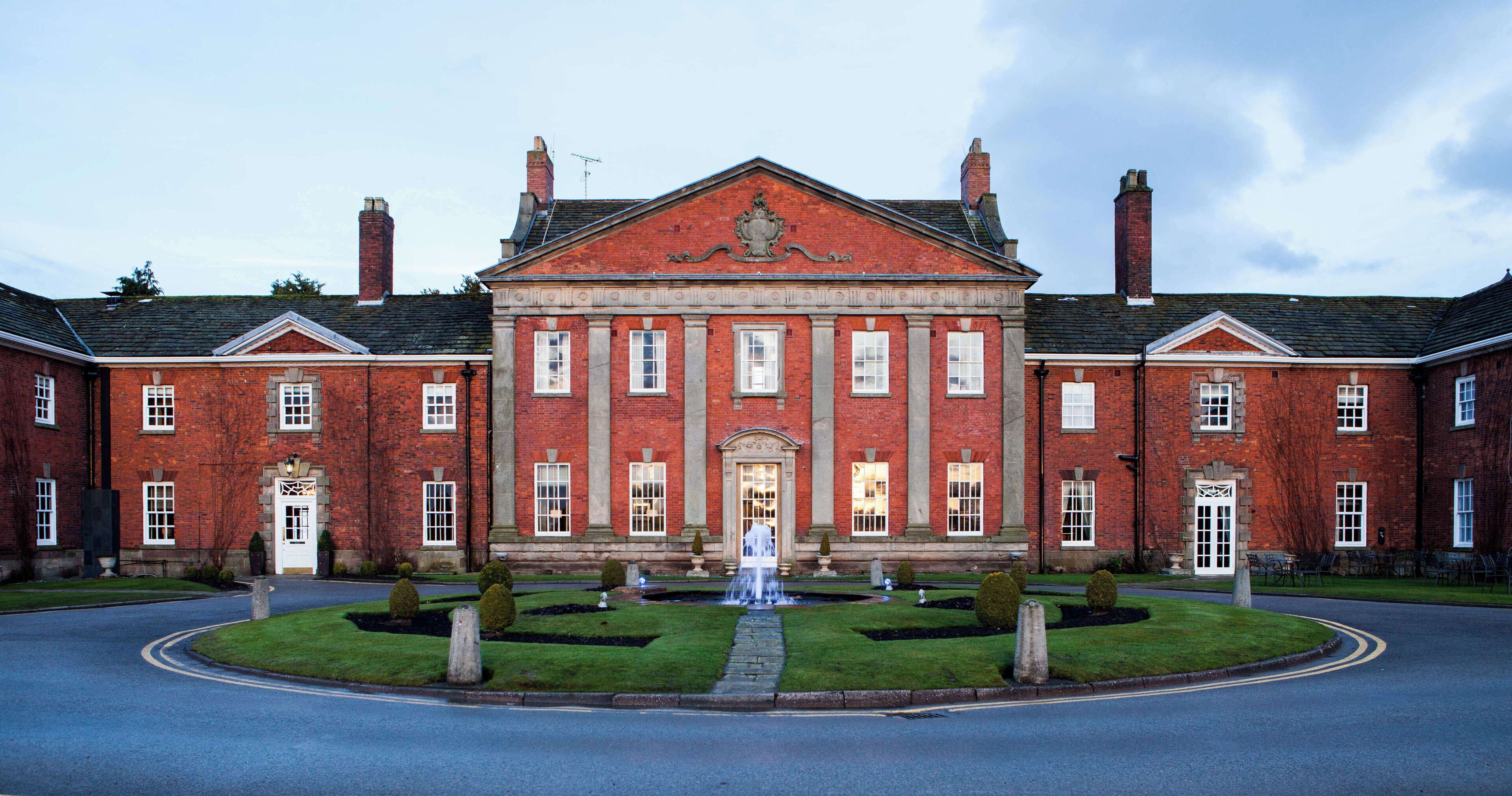 A large red brick building with a symmetrical facade, a central entrance with a decorative pediment, and a well-maintained lawn with a fountain.