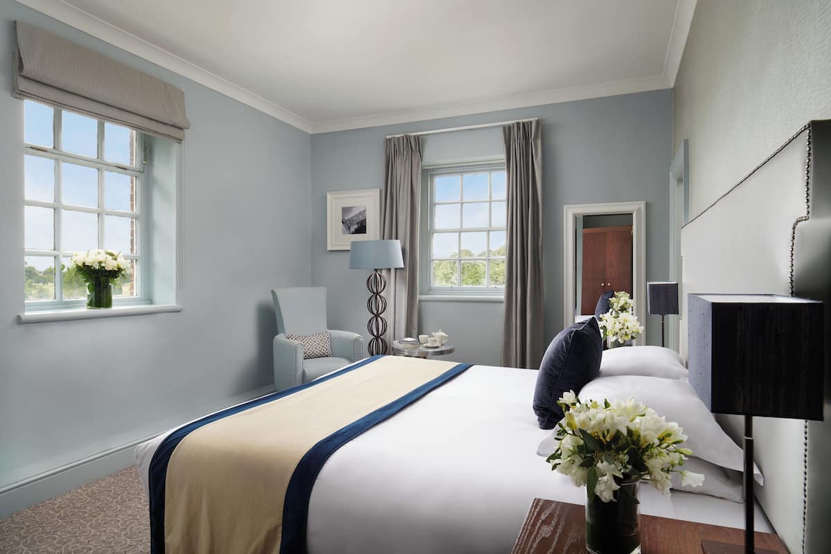 A hotel room with a large bed, two windows with curtains, a bedside table with a lamp, a chair, and a vase of flowers.
