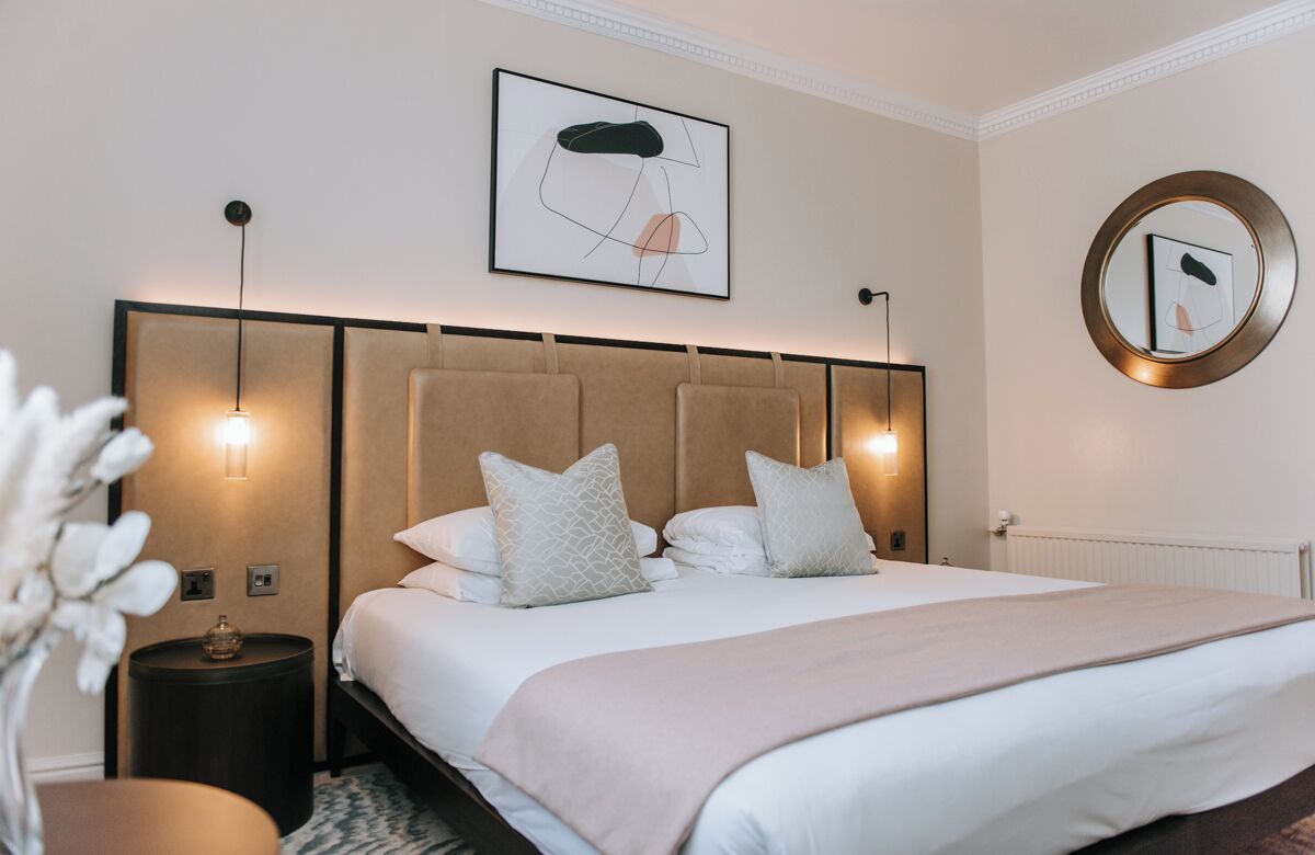 A neatly arranged bedroom with a large bed, bedside lamps, a round mirror, and a framed abstract artwork on the wall.
