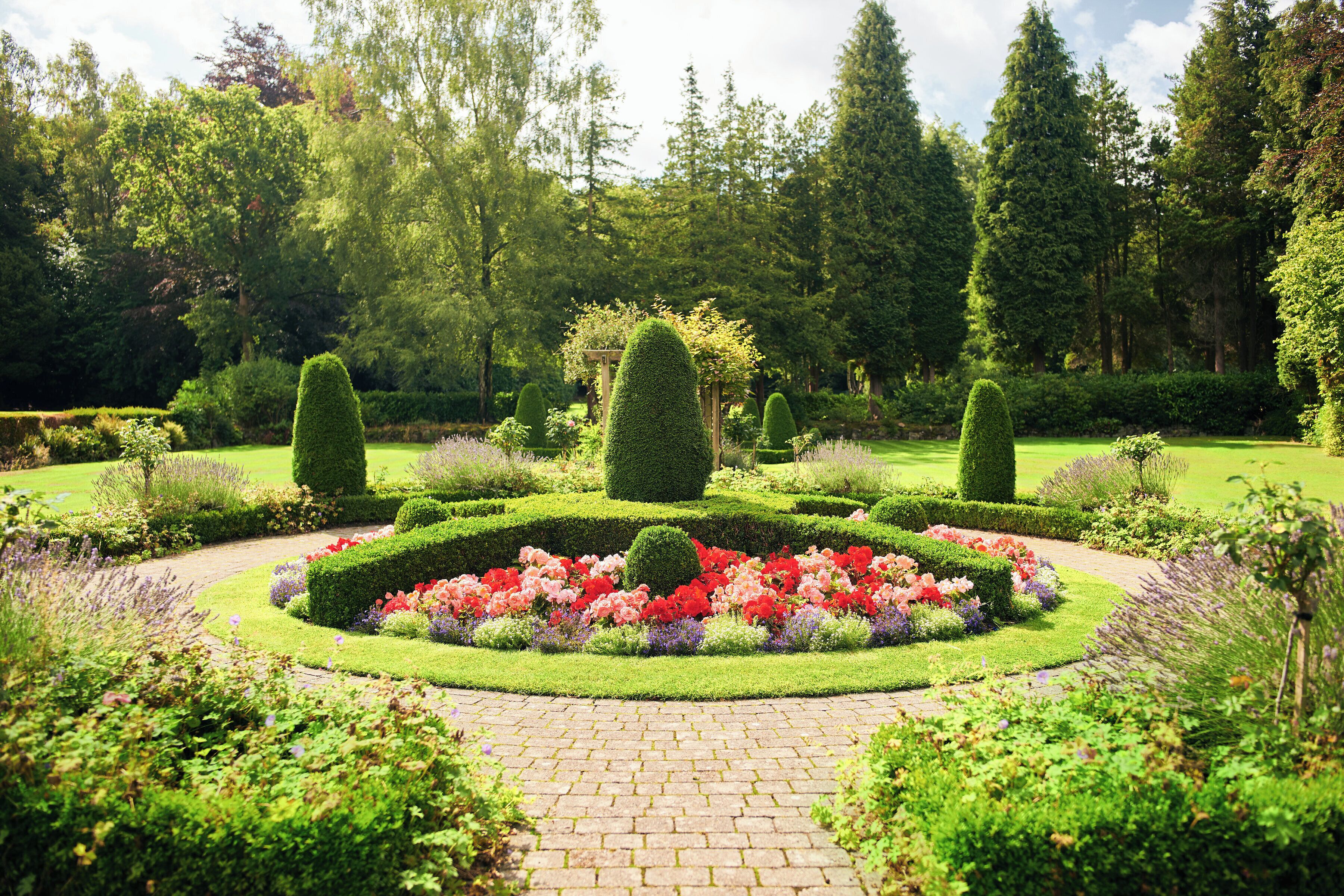A formal garden with a central circular flower bed, trimmed hedges, and a brick pathway.