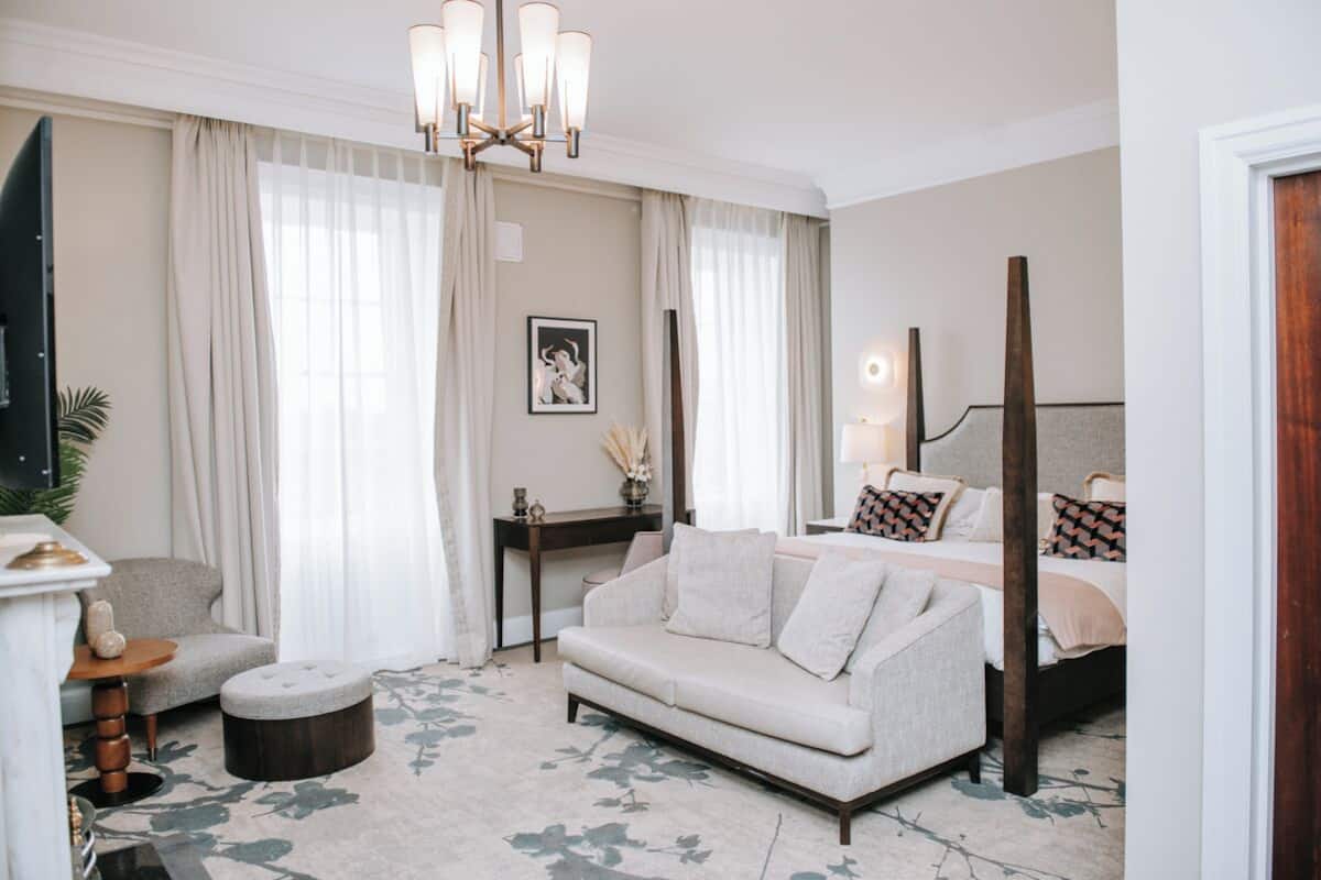 A hotel room with a sofa, a round table, a bed with patterned pillows, a standing lamp, and a large window with sheer curtains.