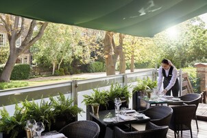 An outdoor dining area with a table set for two, surrounded by wicker chairs, and a waiter attending to the table.