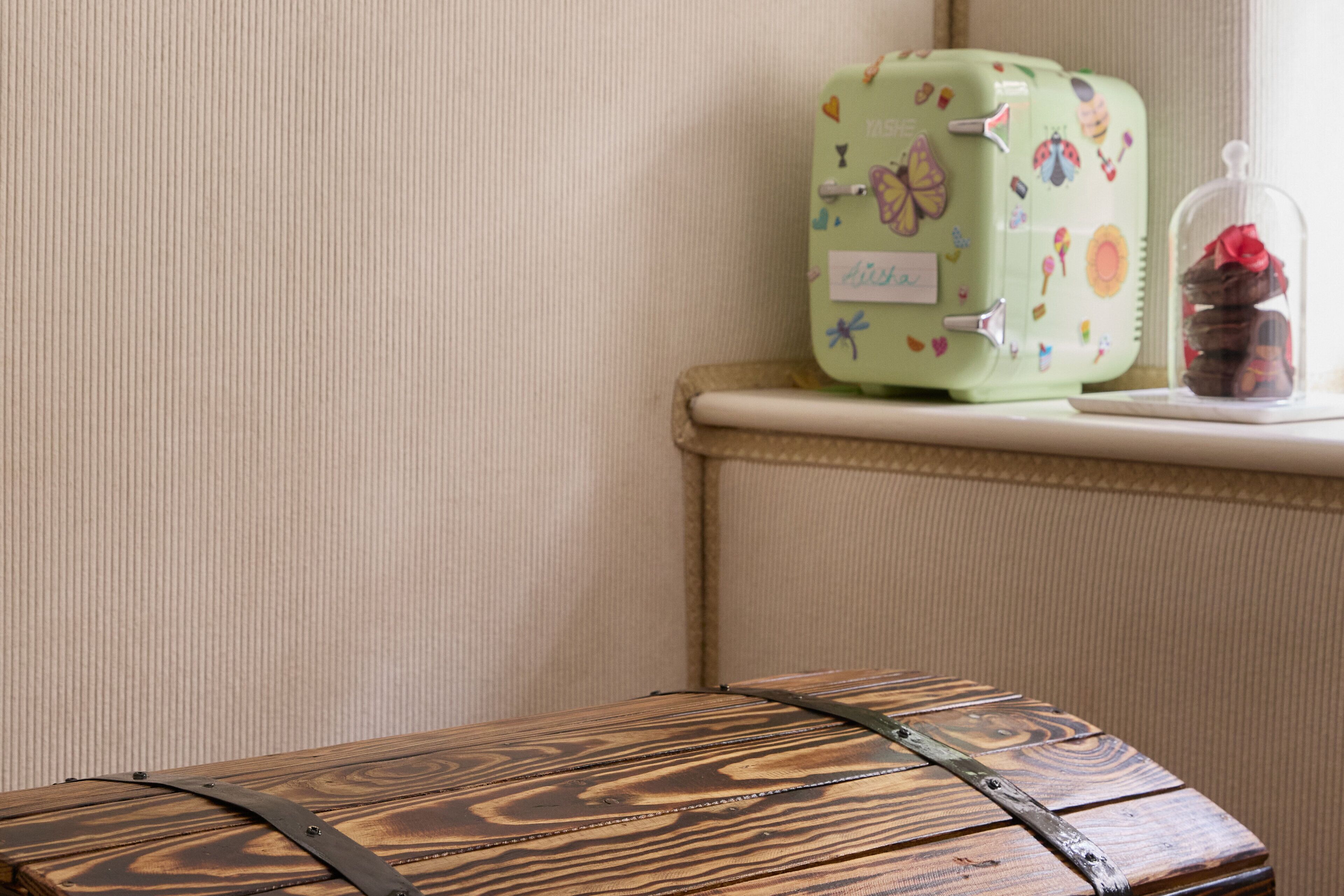 A green suitcase with butterfly stickers on a shelf, a wooden chest with a metal strap, and a glass dome with chocolates.