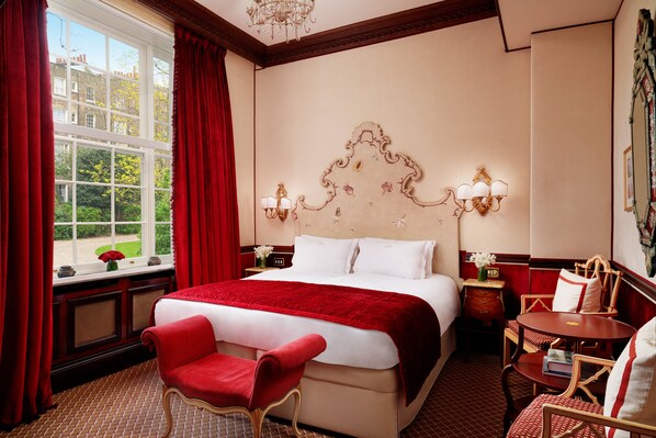 A hotel room with a large bed, a red chair, a small table, and a view of the outside through a window. - The Montague On The Gardens (London)