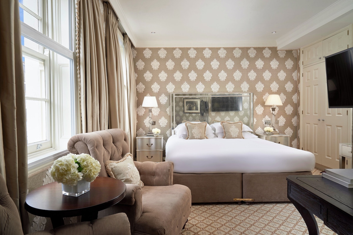 A hotel room with a large bed, a sitting area with a table and chair, a TV, and a window with curtains.