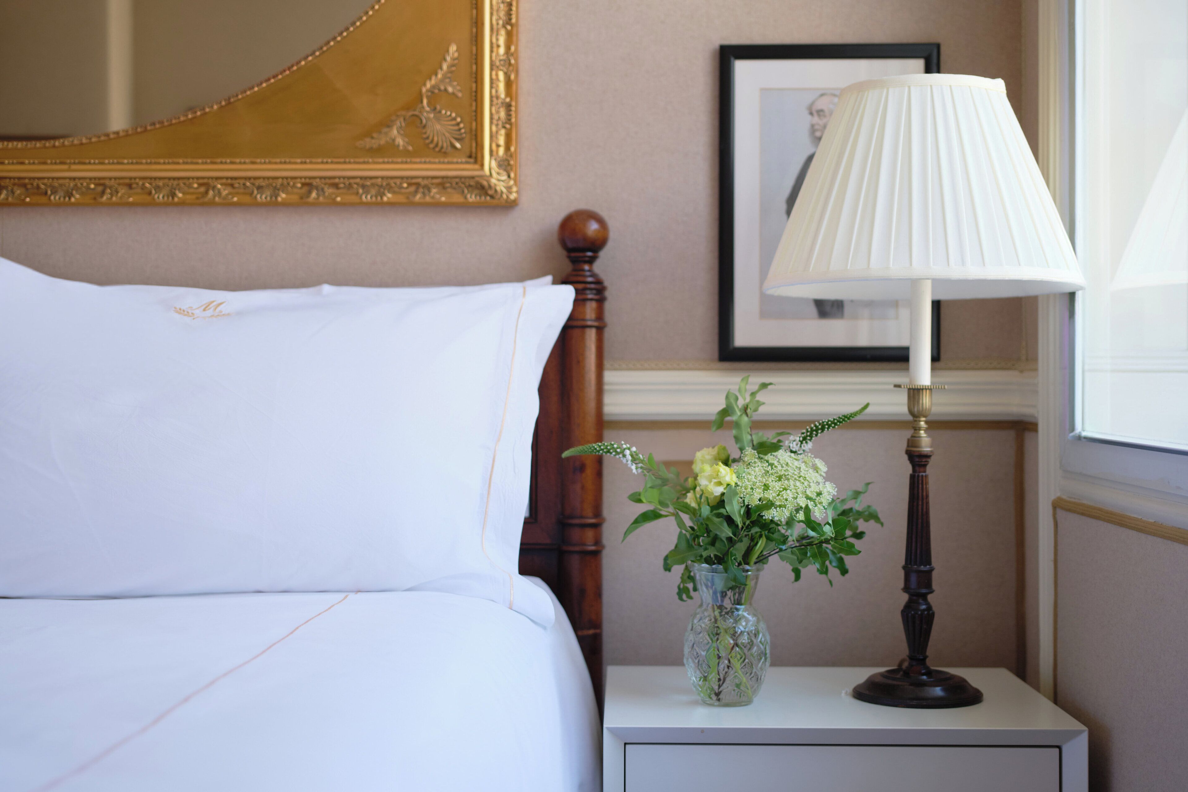 A neatly made bed with white linens, a bedside table with a lamp and a vase of flowers, and a framed picture on the wall.