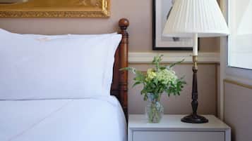 A neatly made bed with white linens, a bedside table with a lamp and a vase of flowers, and a framed picture on the wall.