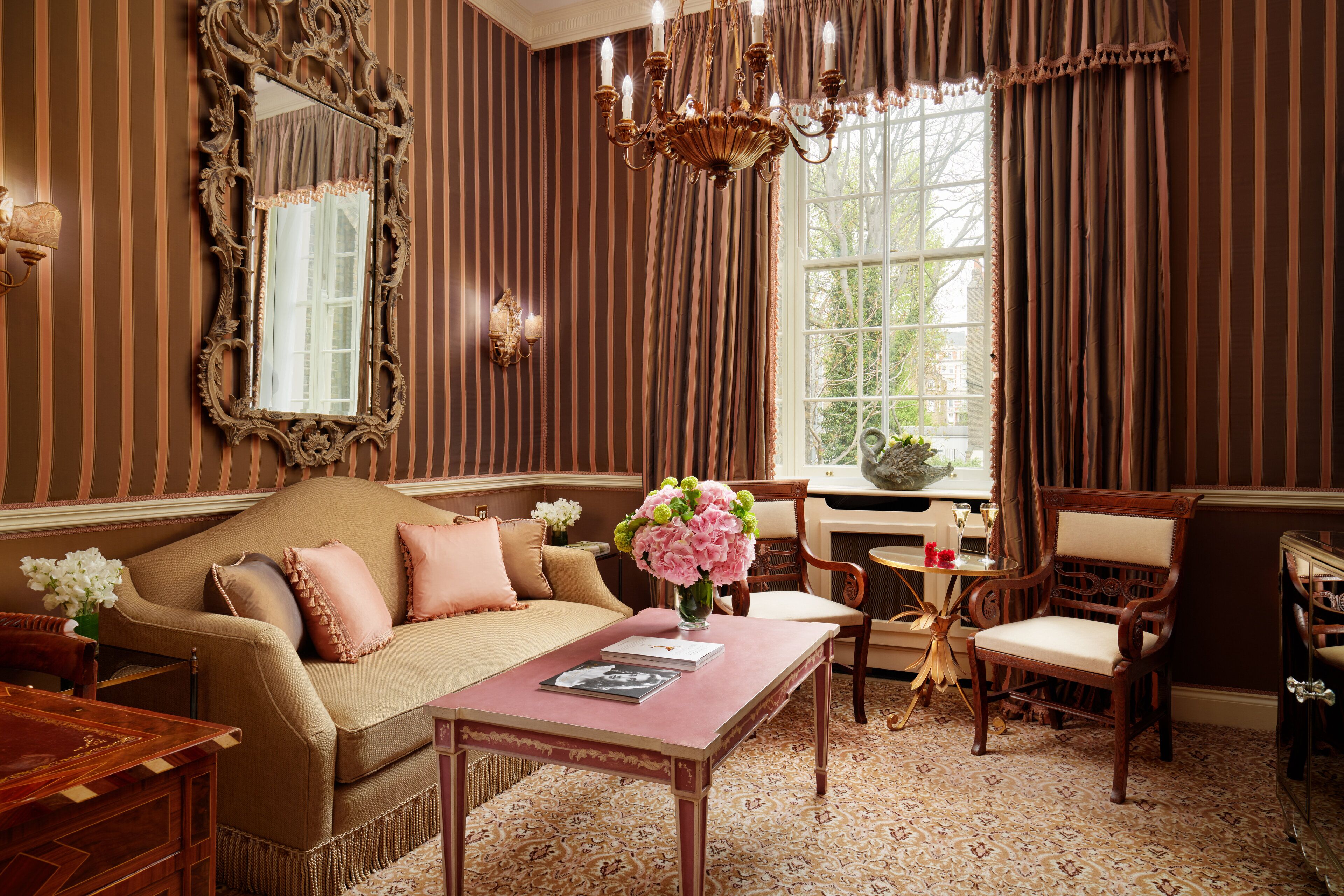 A room with a striped wallpaper, a large mirror, a chandelier, a sofa with cushions, a table with a vase of flowers, and a window with curtains.