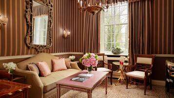 A room with a striped wallpaper, a large mirror, a chandelier, a sofa with cushions, a table with a vase of flowers, and a window with curtains.