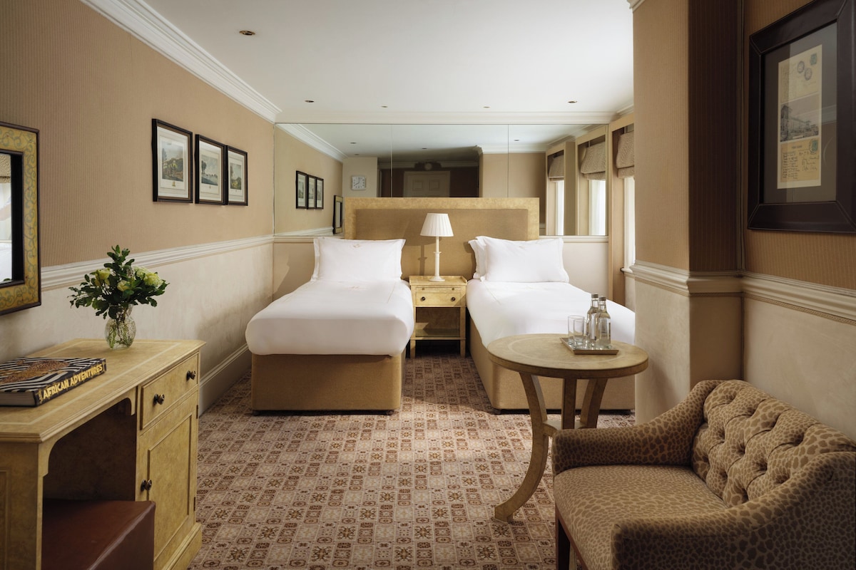 A hotel room with two beds, a small table, a sofa, a nightstand, a lamp, and a vase with flowers.