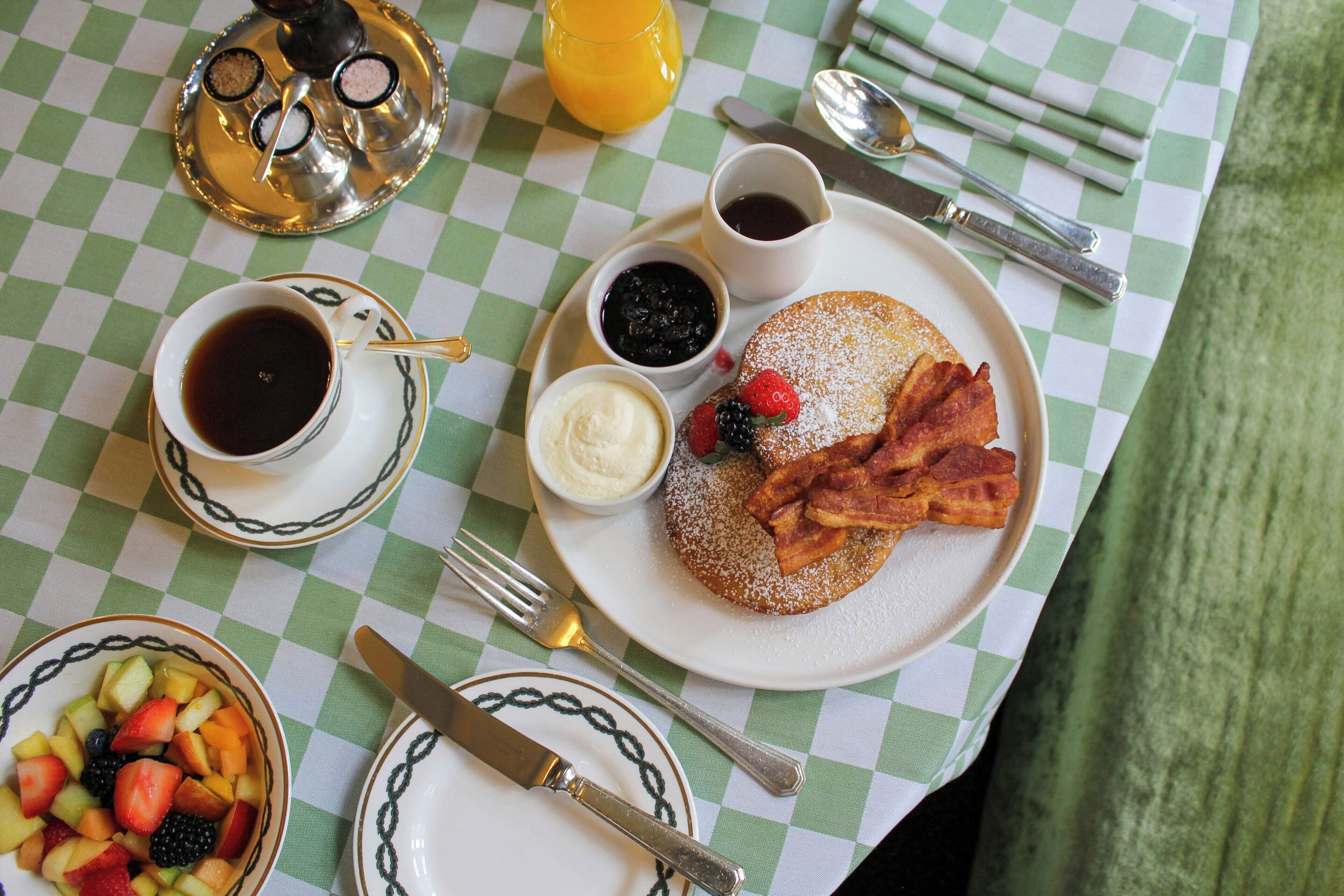 A breakfast table with a checkered tablecloth, a plate of pancakes with bacon and berries, a cup of coffee, a bowl of fruit, and a glass of orange juice.