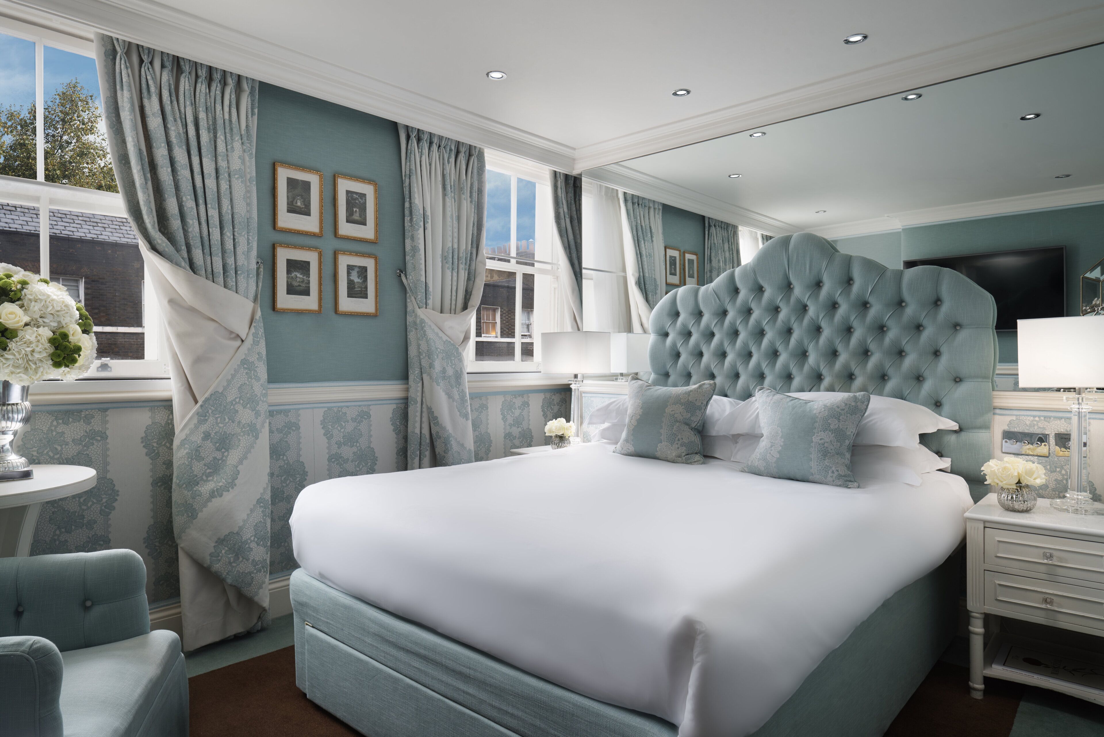 A large bed with a tufted headboard, a bedside table with a lamp, and a view of the outside through a window with curtains.