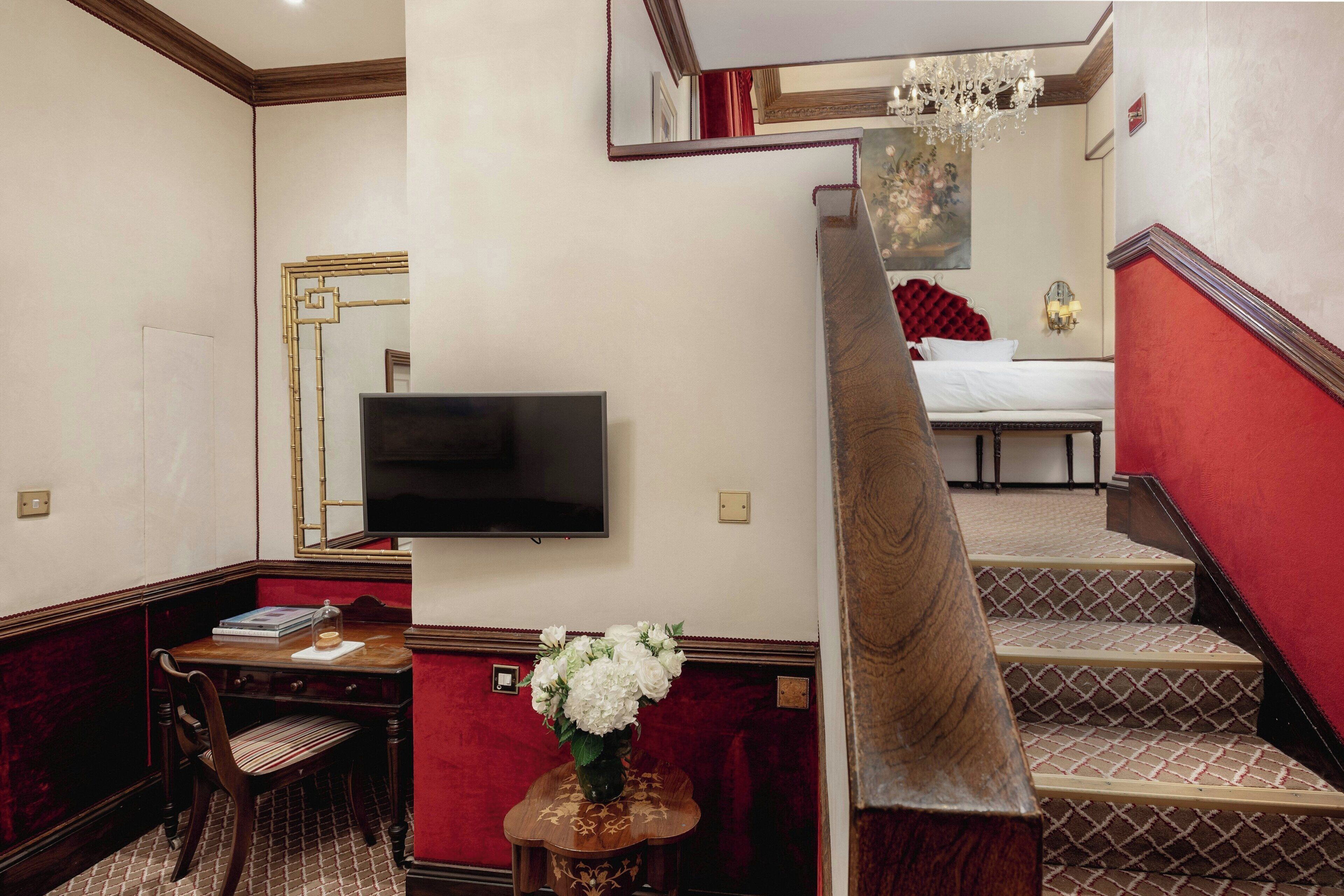 A hotel room with a TV, a dining table with chairs, a vase of flowers, a staircase with red walls, and a chandelier.