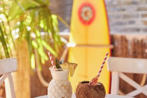 A tropical drink with a straw and a slice of pineapple, a coconut with a straw, and a yellow surfboard in the background.