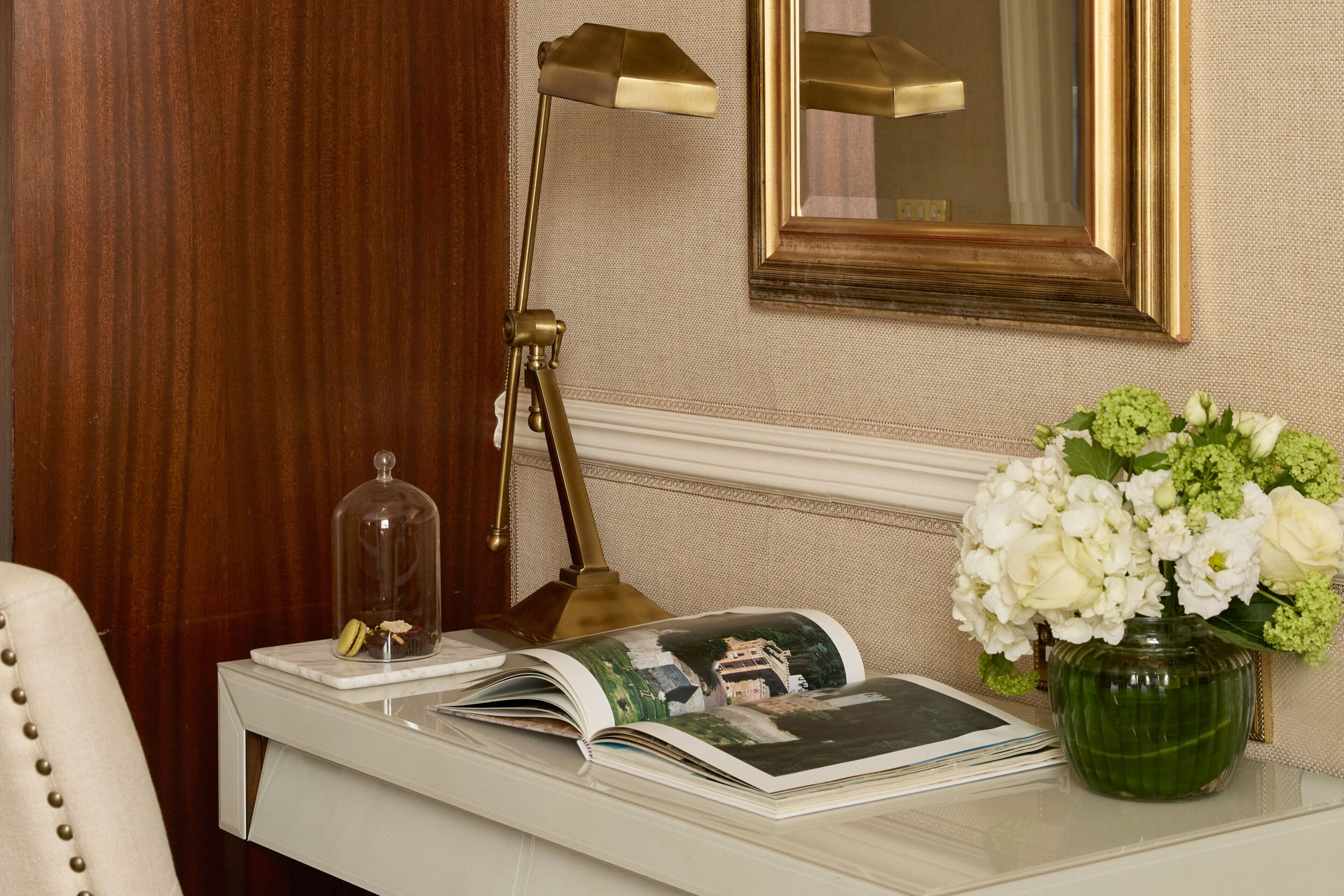 A desk with a lamp, a glass dome, a book, and a vase with flowers.
