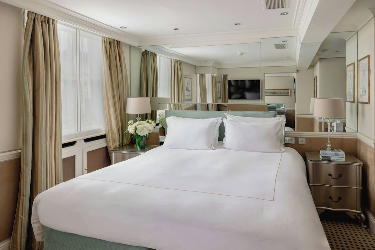 A hotel room with a large bed, a bedside table with a lamp, a mirror, and a window with curtains.