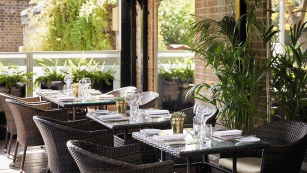 Un espacio de comedor al aire libre con mobiliario de mimbre y plantas en macetas. Las mesas están dispuestas con platos, vasos y cubiertos.