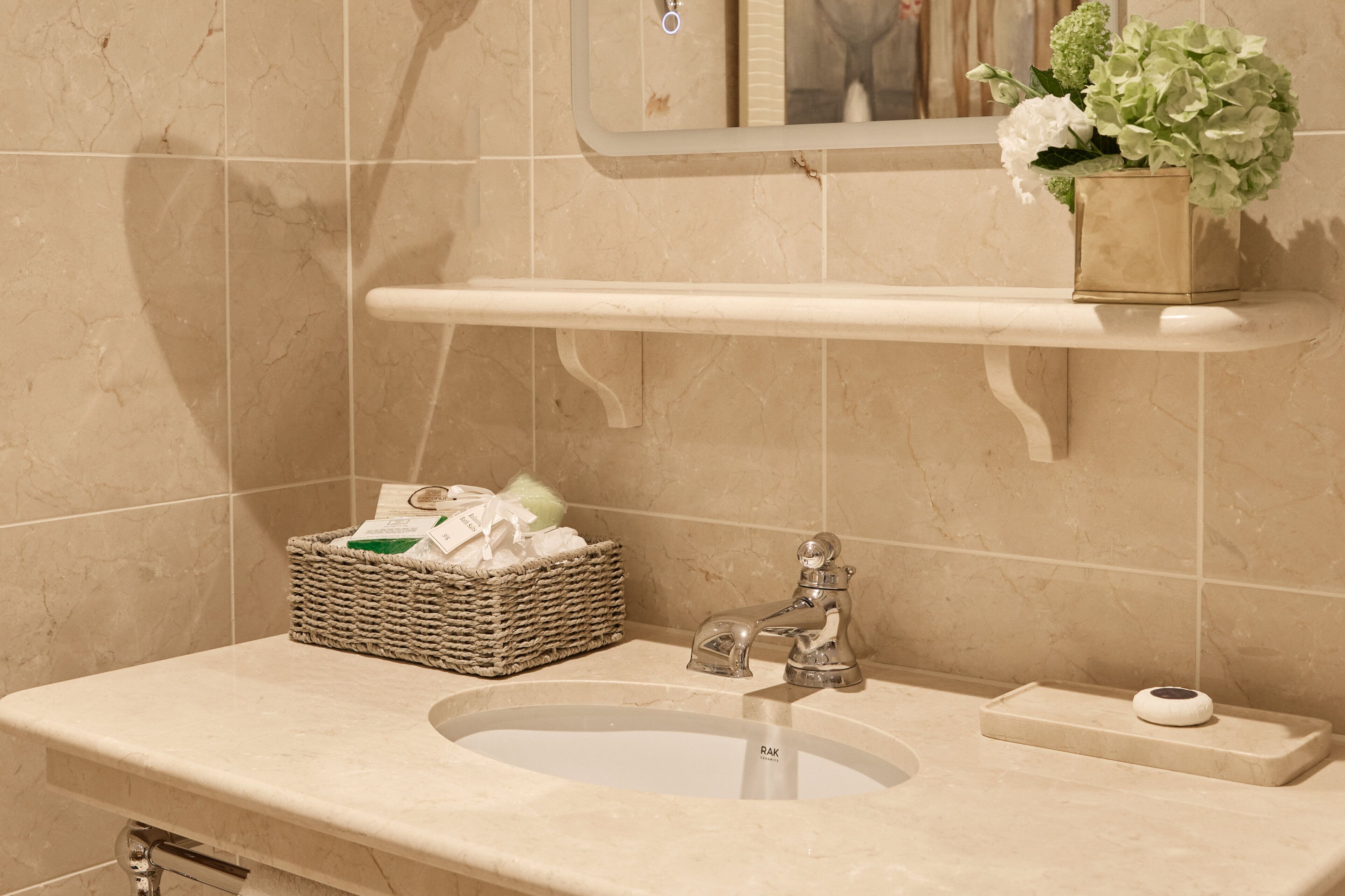 A bathroom with a marble sink, a mirror, a shelf with a potted plant, and a wicker basket with toiletries.