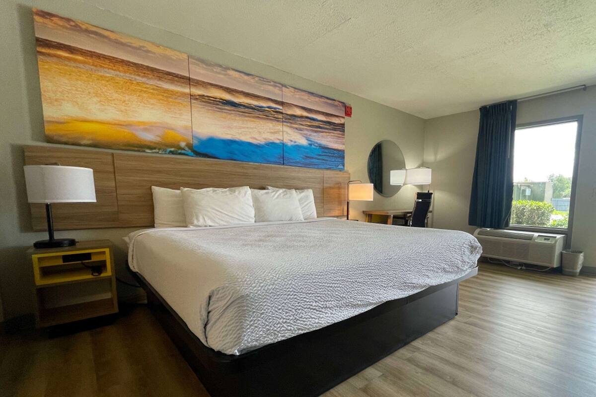 A hotel room with a large bed, a nightstand, a lamp, a mirror, and a window with curtains.