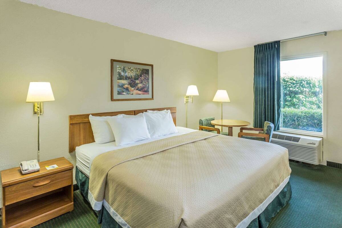 A hotel room with a bed, bedside tables, a desk, a chair, a window with curtains, and a painting on the wall.