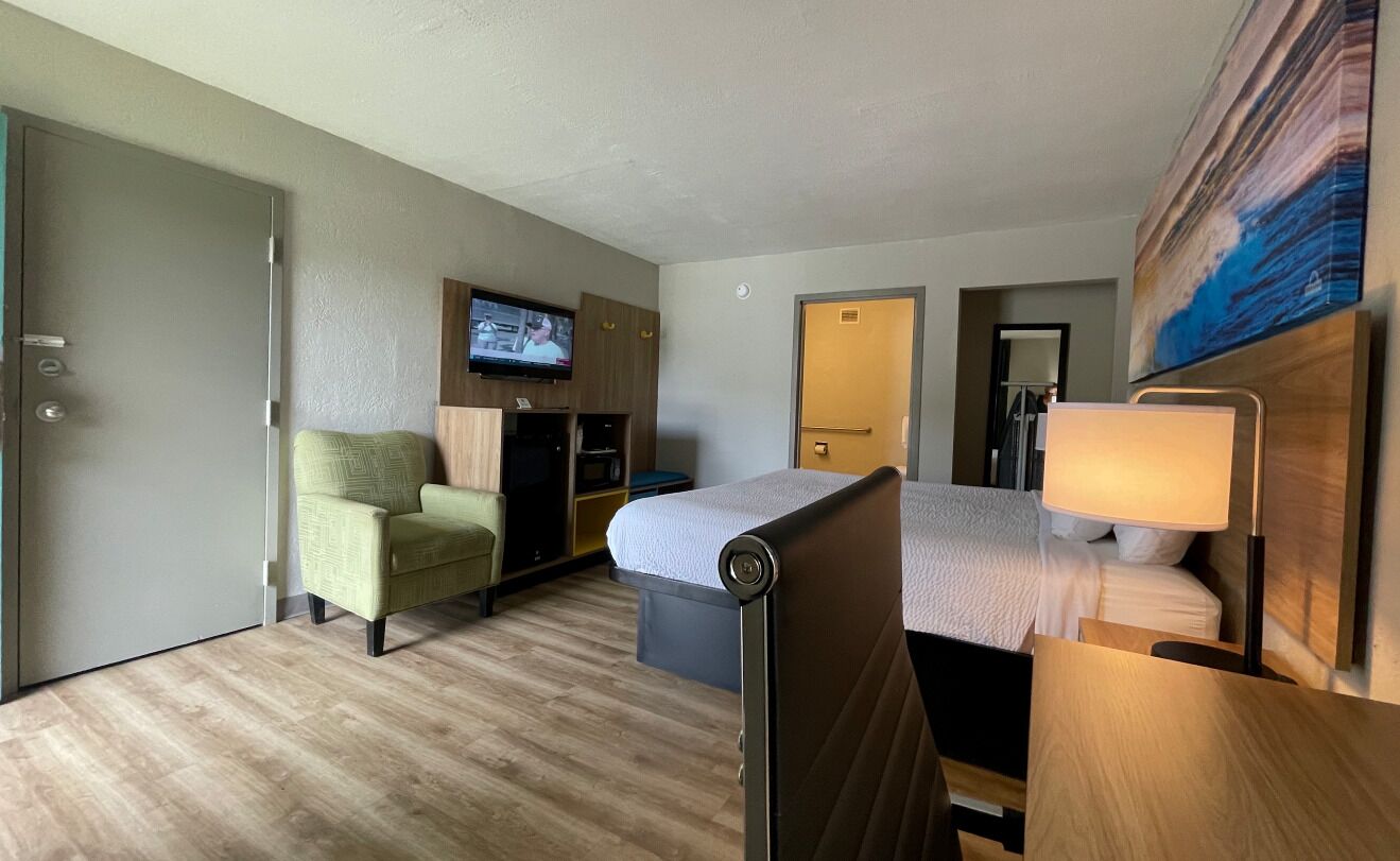 A hotel room with a bed, a desk, a chair, a TV, and a painting on the wall.