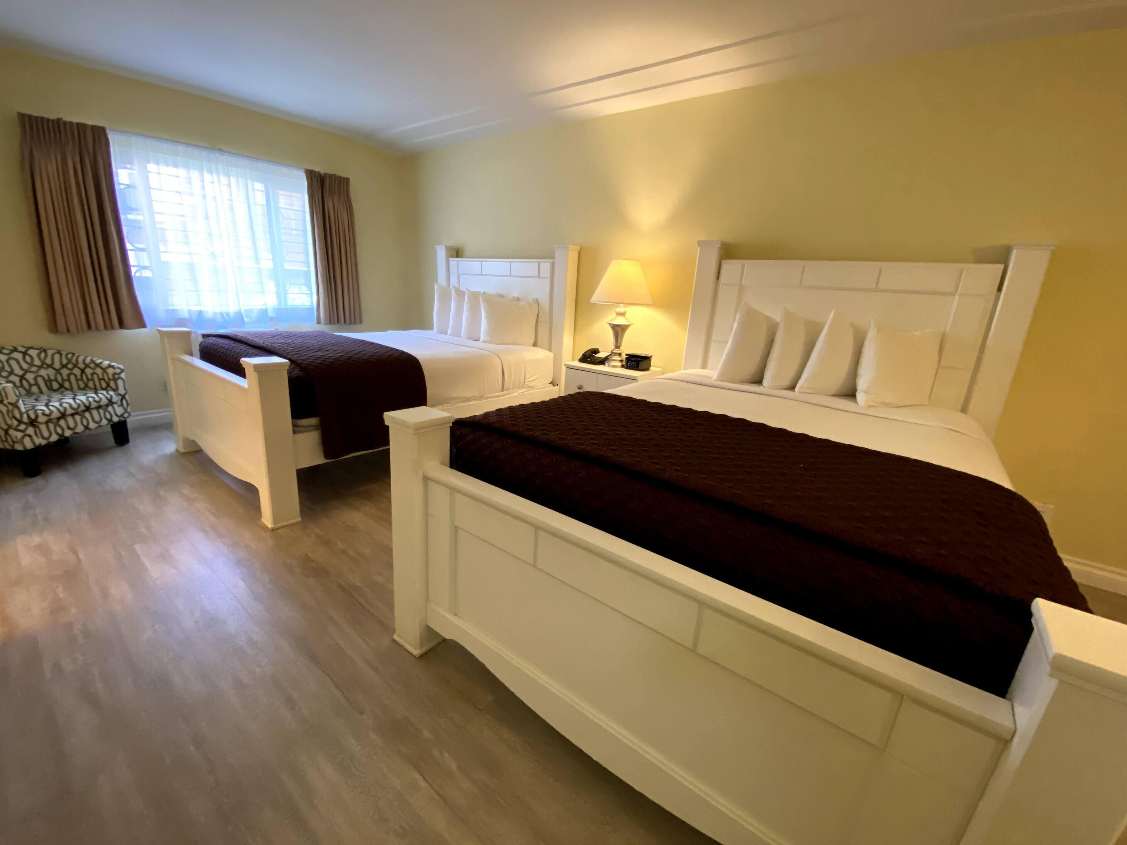 Une chambre d’hôtel avec deux lits simples, une chaise, une table de chevet avec une lampe et une fenêtre avec des rideaux.