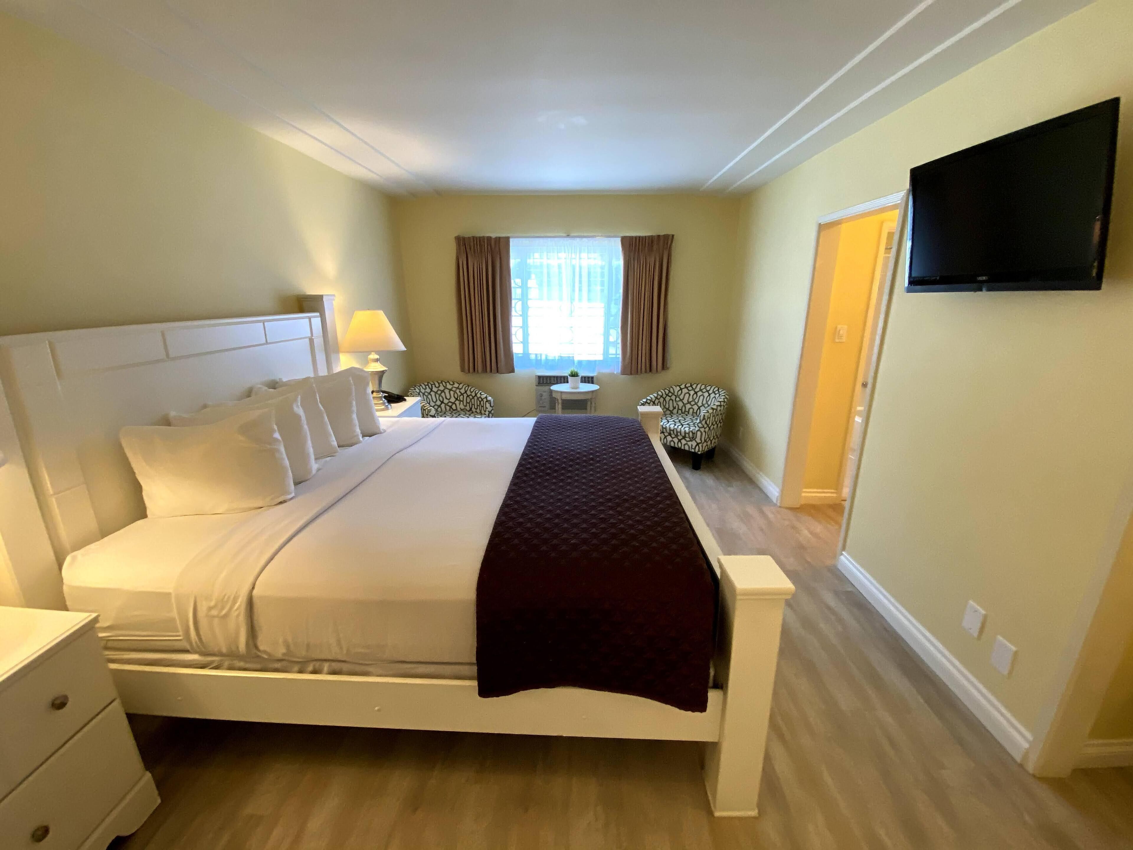 A hotel room with a large bed, a bedside table, a chair, a TV mounted on the wall, and a window with curtains.