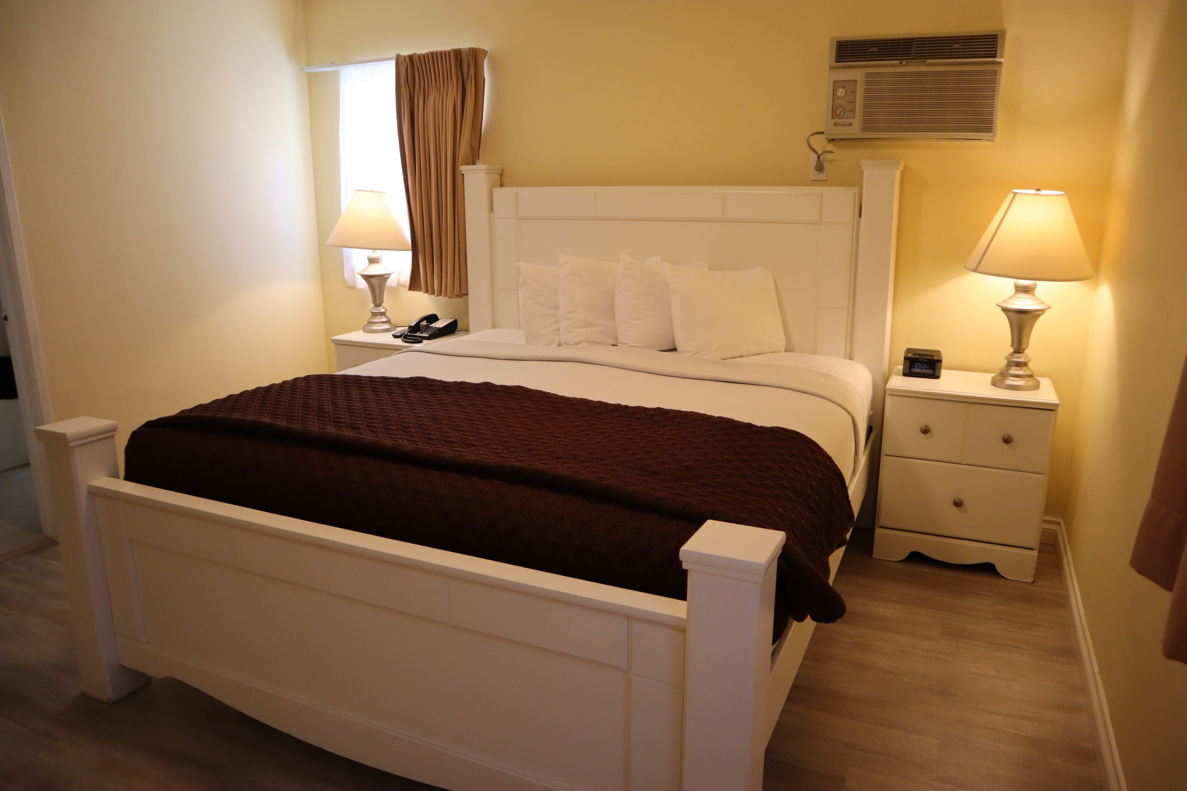 A neatly arranged bedroom with a white bed, a bedside table with a lamp, and an air conditioning unit on the wall.