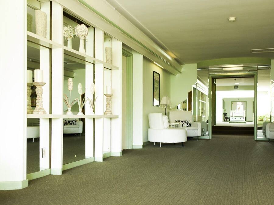 A spacious hallway with a carpeted floor, white walls, and a series of white chairs and tables.