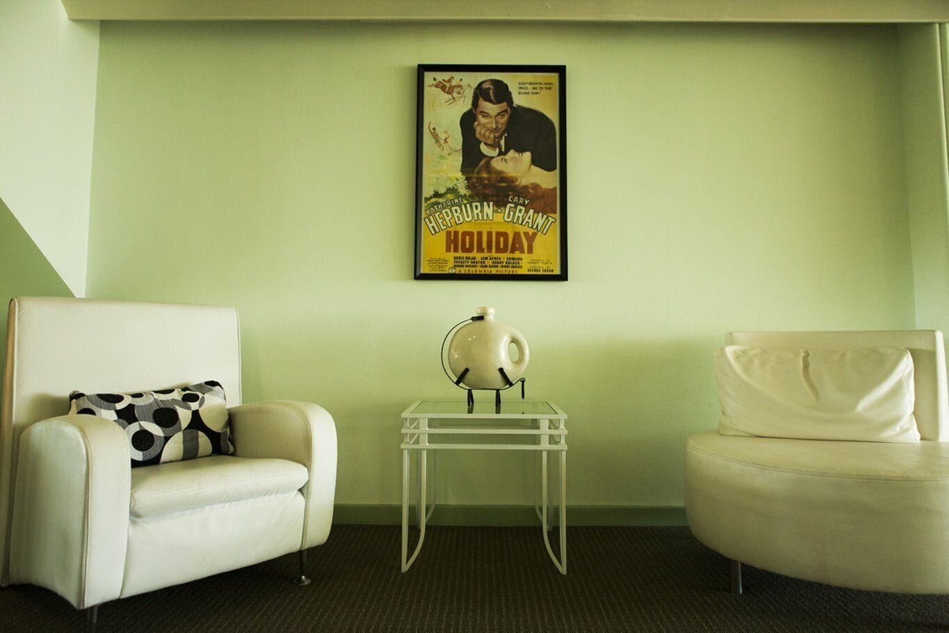 A living room with two white armchairs, one with a patterned pillow, and a small table with a globe. A vintage movie poster hangs on the wall.