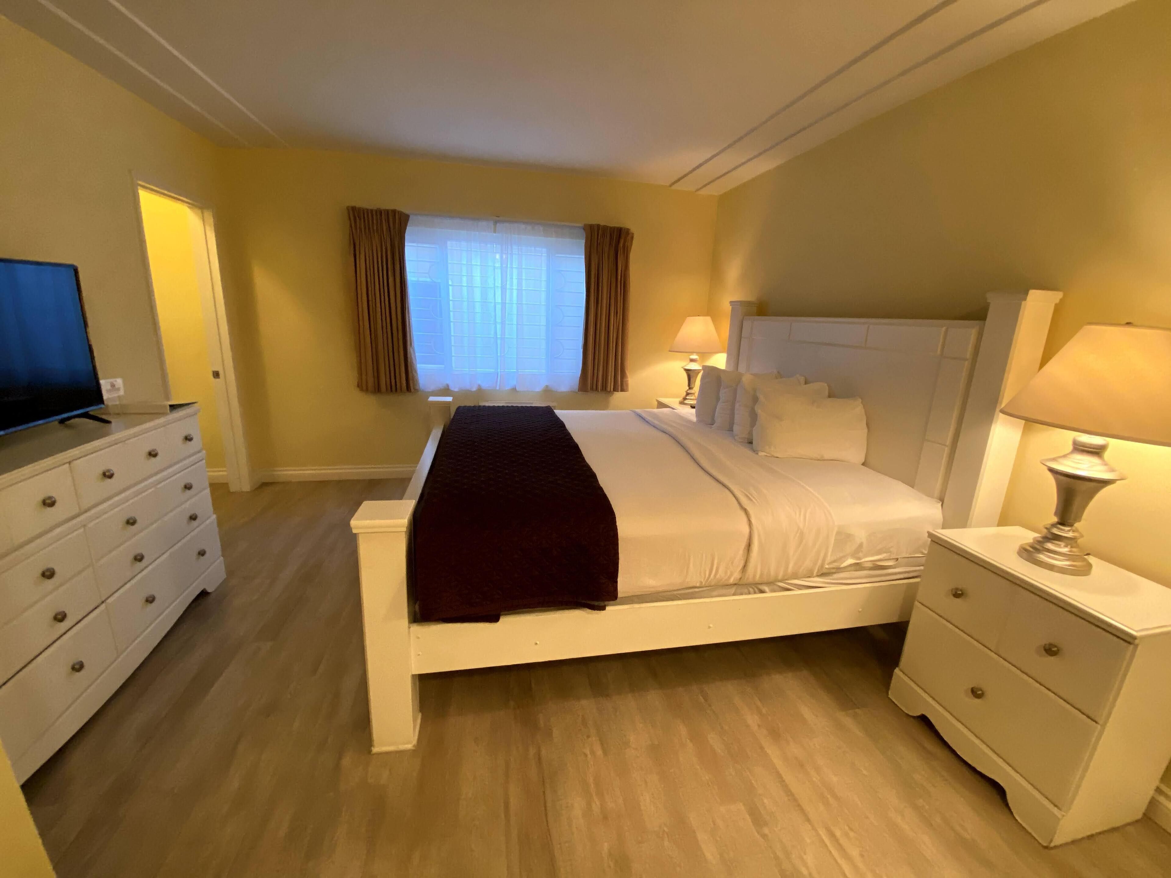 Habitación de hotel con una cama grande, dos mesitas de noche con lámparas, un televisor en una cómoda y una ventana con cortinas.