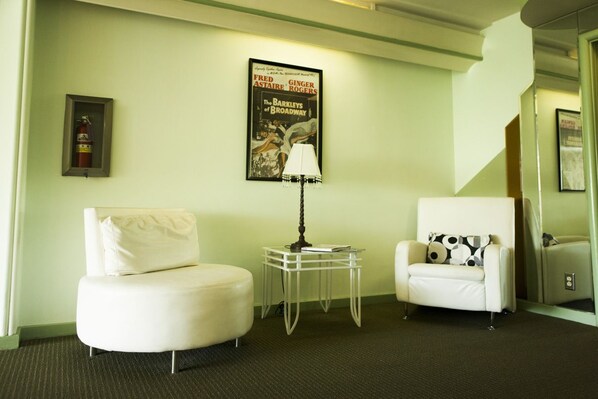 Habitación de hotel con un sillón blanco, un sofá redondo blanco, una mesita de noche con una lámpara y un cartel en la pared.