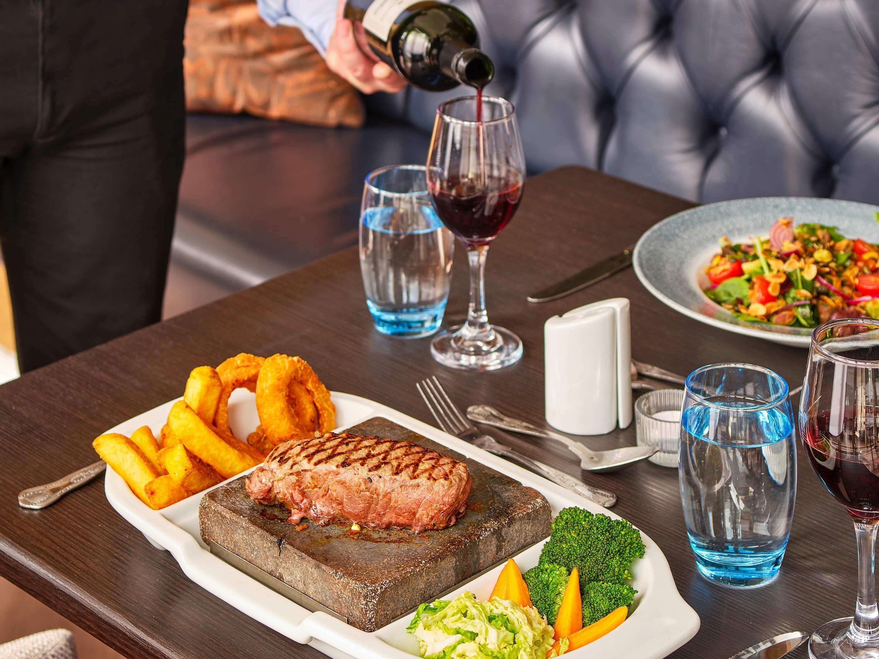 A dining table with a steak, onion rings, and vegetables, a bottle of red wine, and a plate of salad.